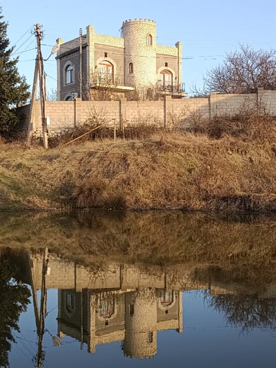 A charming building with a turret sits atop a grassy embankment, reflected in a still body of water. The structure features light-colored stone accents against gray walls, with balconies and arched windows. A stone wall runs along the property line. Dry grass and shrubbery cover the bank, and a utility pole stands to the left. Trees, both bare and evergreen, frame the scene under a clear sky. The water mirrors the entire setting in an almost symmetrical composition.