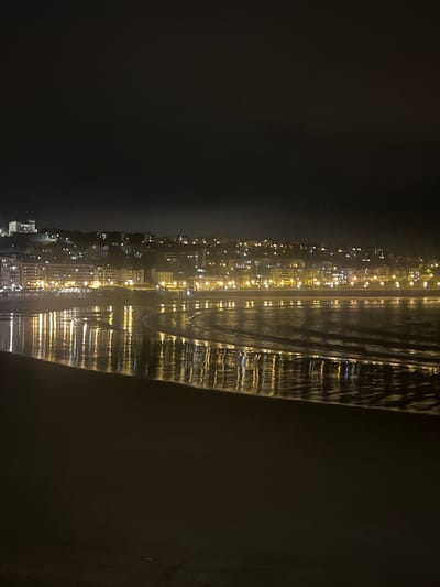 The image depicts a nighttime scene of the La Concha beach in San Sebastián, Spain. The foreground is a dark, sandy beach. Beyond the beach, the calm waters of the bay reflect the lights of the city, which stretches along the coastline and up the hills in the background. The city is illuminated with numerous bright lights, indicating a vibrant urban area at night. The sky is dark, with a slight hazy quality that diffuses the city lights. The reflections on the water create a shimmering, almost abstract pattern of light. There are no people or distinct objects visible in the image, creating a peaceful and somewhat ethereal atmosphere.