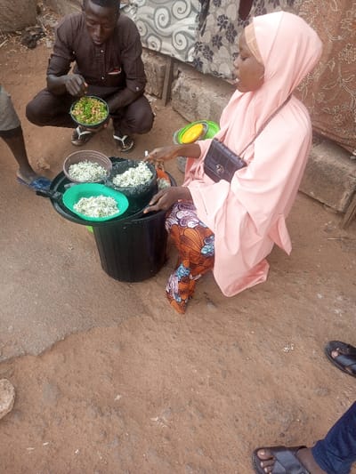 This image captures an outdoor food preparation or serving scene in Birnin Kebbi, Nigeria. In the foreground, a woman wearing a light pink hijab and abaya, with patterned trousers, is seated beside a black plastic bucket acting as a low table. On a black plastic tray atop the bucket, three bowls contain a granular food item, likely rice mixed with green vegetables or herbs. The woman's right hand, holding a utensil, is positioned near the food, suggesting she is serving or mixing. A black handbag with a chain strap is slung over her shoulder. To her left, a man squats, dressed in a brown long-sleeved tunic and flip-flops. He holds a green bowl filled with a similar granular dish, with his fingers inside the bowl. The ground is predominantly reddish-brown earth, with sections of concrete and small stones. In the background, two pieces of patterned fabric, one with gray spirals and another with brown florals, hang as a makeshift wall or shade, supported by visible bricks at their base. The feet of at least two other individuals are partially visible: one wearing blue slide sandals on the far left, and another in black leather sandals on the bottom right.