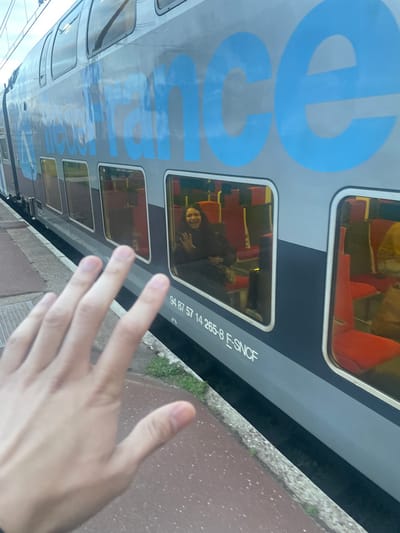 An outdoor, eye-level photograph captures a moment at a train station platform in Plaisir, France. In the foreground, the right hand of an unseen individual is extended towards a stationary double-decker train, palm open with fingers slightly spread. The train, a gray "E-SNCF" model with large blue "France" lettering along its side, is visible from its mid-section to its roofline. Through one of the train's lower windows, a female passenger with dark hair and a dark jacket is visible, seated on a red-upholstered chair, smiling and waving her right hand towards the photographer. Other red seats are visible within the train's brightly lit interior. Below the windows, the train bears the identification number "94 87 57 14 265-8 E-SNCF". The platform, composed of reddish-brown paving stones with a concrete edge, runs along the left side of the train. A metal grate is embedded in the platform to the left. A narrow strip of green grass is visible between the platform's edge and the train's lower body. The sky, appearing overcast, is reflected in the train's windows and upper metallic surface.