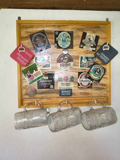 This image displays a collection of beer-related memorabilia, primarily beer labels and coasters, mounted on a light-colored, rustic wooden board. Three empty, clear glass beer mugs are hanging vertically on the right side of the board. The beer labels vary in design and include text in German, such as "TRINKEN IST FERTIG!", "DUNKEL", "LAGERBIER HELL", "Helles Landbier", and "SCHWARZBIER". There are also coasters with text like "HEUTE: Betreutes Trinken" and "TANNEN ZÄPFLE VOM HOCHSCHWARZWALD". The overall scene suggests a casual, possibly domestic setting, like a home bar or a collection displayed on a wall in Germany. The items are arranged somewhat randomly, with the mugs adding a three-dimensional element to the display. The lighting is even, indicating an indoor setting.