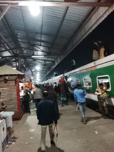 A bustling train station platform under a large metal roof. Crowds of people are walking along the platform beside a green and white train. Some individuals are boarding the train, while others stand or walk nearby. A vendor offers food to passengers. Several people are on top of the train. A small kiosk stands to the left of the frame. The scene appears to be dimly lit.