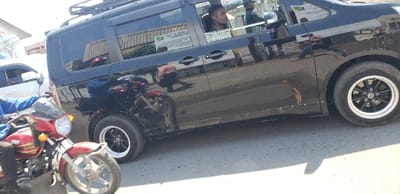 The image shows a black van parked on a street in Rubavu, Rwanda. The van has a luggage rack on its roof and is parked on the right side of the road. A red and silver motorcycle is in the foreground. There is another white vehicle partially visible in the background.  The man in the driver’s seat of the van is looking forward while holding the steering wheel. The motorcycle rider is wearing blue clothing. The sky is clear. The location is likely a residential street in Rubavu, Rwanda, based on the coordinates provided.