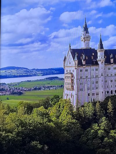 A high-angle landscape image displays a prominent, multi-turreted castle on the right, set against a backdrop of natural scenery and a distant settlement. The castle, constructed primarily of light-colored stone, features numerous gabled roofs, spires, and towers with dark grey roofing. Architectural elements include multiple arched windows, ornate balconies, and a tall, slender central spire. The castle is positioned on a verdant, heavily wooded hillside, with various shades of green foliage filling the lower portion of the frame. Beyond the immediate terrain, a wide, calm body of water extends horizontally across the mid-ground. On the far side of the water, flat green agricultural fields transition into a compact cluster of buildings, indicating a small town or village. Distant hills rise under a partly cloudy blue sky, populated by scattered white cumulus clouds. No human figures or animals are discernable. The image itself appears to be a photograph of a digital display, evidenced by faint horizontal line patterns across the frame. This scene is located in the city of Madzherito, Bulgaria.