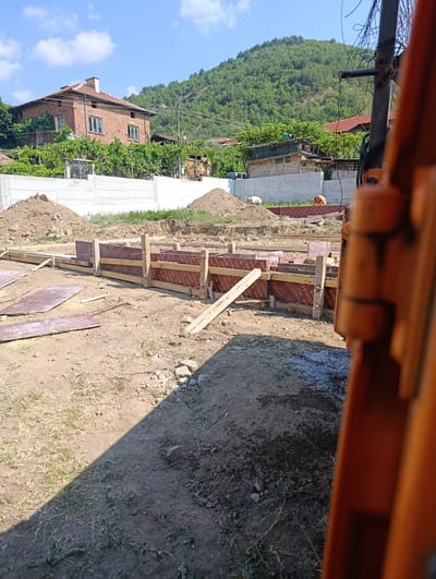 The image depicts an outdoor construction site under a bright, sunny sky, suggesting it is daytime. The scene is a building foundation being prepared. Wooden forms, lined with reddish-brown material, are set up in rectangular shapes on the excavated earth, ready for concrete. Several piles of dirt are visible around the foundation area. In the background, a residential house made of brick with a tiled roof is situated, surrounded by greenery, with a lush, tree-covered hill rising behind it. A concrete wall runs along the front of the property. Two individuals are faintly visible in the distance, seemingly working on the site. The foreground is dominated by the construction elements and the dusty ground. Some pieces of plywood and rebar are also scattered around the foundation. The overall impression is one of ongoing construction in a rural or semi-rural setting. Visible text on the wooden forms reads "GTC FORMWORK" and "GRADIPLEX".