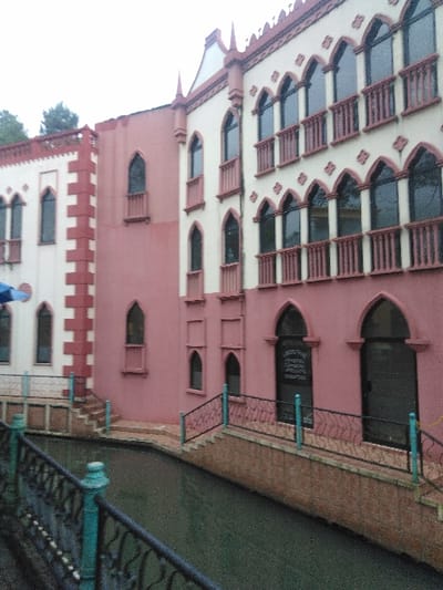 The image displays a multi-story building complex with Venetian Gothic-inspired architecture, situated alongside a canal in Ciudad de San José, Costa Rica. The buildings are predominantly colored in light pink/mauve, accented with off-white/cream trim. The taller structure on the right features multiple levels of narrow, tall windows with pointed Gothic arches; some windows on the upper levels have small, light-colored balconettes with short balustrades. Intricate decorative elements, including pointed finials, adorn the roofline and upper facades. The attached shorter building on the left exhibits similar architectural characteristics, with prominent off-white block-like trim. A narrow canal, filled with dark green water, runs horizontally across the foreground, its banks constructed from reddish-brown brick-like material. Steps descend from the building's base to pathways alongside the canal. Black metal railings with decorative scrollwork and turquoise posts border these pathways and steps. The sky above is overcast. No individuals are present in the scene.