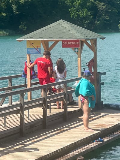 A lifeguard stands on a wooden dock next to a lifebuoy and a life jacket. A woman in a bathing suit and a turquoise towel wrapped around her shoulders, is also on the dock. In the background, a man in a red shirt with "GELBĖTOJAS" printed on the back, stands with his hand on a wooden post. A woman in a white shirt and shorts is standing next to him. A sign above them reads "GELBĖTOJAI" which means lifeguards in Lithuanian. Another sign depicts a person diving and states "DRAUDŽIAMA NARDYTI," meaning diving is forbidden. The scene appears to be at a lake or river in Vilnius, Lithuania, given the signage. The water is a vibrant blue-green.