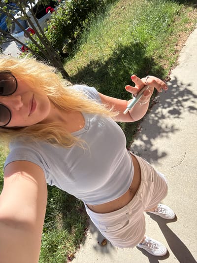 A young blonde woman is captured in a selfie, holding a cylindrical object in her right hand, which appears to be an e-cigarette. She is wearing a light blue ribbed crop top that reveals her midriff, featuring a belly button piercing. Her lower half is adorned with distressed white denim shorts, and she has on white athletic shoes. The woman has on dark sunglasses and her blonde hair cascades around her shoulders.

The setting is outdoors, on a sunny day, indicated by the strong shadows cast on the concrete path and grass. The time of day is likely afternoon due to the angle of the sun. The environment includes a patch of green grass with scattered fallen petals, a concrete walkway, and in the background, glimpses of greenery and a car, suggesting a residential or park-like area. The overall mood is relaxed and casual. No visible text is present in the image.