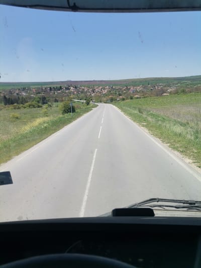 The image depicts a view from inside a vehicle, likely a car or truck, driving on a paved road leading into the city of General Kiselovo, Bulgaria. The road is gray with a white center line marking. On either side of the road, there is vegetation, appearing as grass and some scrubby trees. In the distance, a village or town is visible, with what appear to be buildings and trees scattered throughout. The sky is clear and blue. The perspective is from the driver's seat, looking through the windshield, with parts of the vehicle's dashboard visible at the bottom of the frame. There are no people visible in the image.