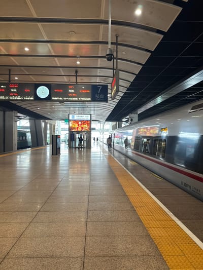 The image depicts the interior of a modern train station platform, specifically Platform 7, located in Fengtai District, China. The platform floor is paved with light-colored, square granite tiles, exhibiting a polished sheen that reflects overhead lighting and nearby subjects. A yellow tactile warning strip with raised dots borders the tracks, paralleled by a solid white line.
On the right, a silver and white high-speed train, identified by "ZE 220711" and "三等座" (Third Class Seat) text on its side, is stopped at the platform. Its windows reflect the station's interior. One individual is seen walking away from the camera along the platform adjacent to this train, while another person's reflection is visible further down the platform.
Above the platform, a large digital display board shows red characters, likely indicating train schedules or destinations, alongside a white analog clock. The designation "7 站台" (Platform 7) is clearly visible. A dark, dome-shaped security camera is mounted from the ceiling structure, monitoring the platform. The ceiling features a curved design with alternating light-colored acoustic panels and dark grid sections, interspersed with recessed lighting fixtures.
In the middle ground, on the left side of the platform, a vertical digital screen displays a vibrant red and gold advertisement, possibly for "光明电器" (Guangming Electric). Several individuals are visible in the distance beyond this screen, appearing to walk or stand. Further left in the background, a portion of a green train is visible on an adjacent track. The station's exterior appears bright through large, distant windows.