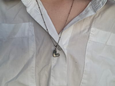 The image is a close-up, overhead shot of a person wearing an open white button-down shirt. A small, silver heart-shaped charm is attached to the shirt, just below the collar. A delicate silver chain hangs from the charm, disappearing down into the neckline of the shirt. The fabric of the shirt is creased and appears to be slightly wrinkled. There are two chest pockets visible on the shirt, one on the left side and one on the right. The background is a plain white surface, likely a wall or a bed. The lighting is even, suggesting it was taken indoors during the day. The image conveys a sense of casualness and personal style. There is no visible text in the image. The location context is Ivatsevichy, Belarus.