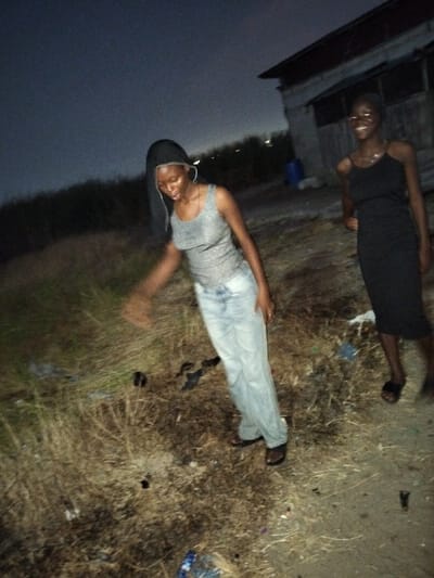 Two young women are standing outside at night. One is wearing a gray tank top and loose jeans. Her head is covered with a dark cloth. The other woman is wearing a black dress. They are standing on an area covered in dried grass, with some trash scattered around. A simple building is in the background. The image has a dark, grainy quality, indicating low light conditions.