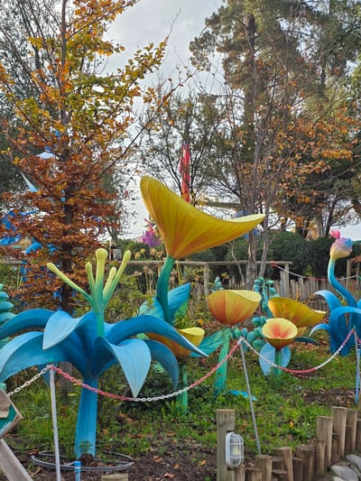 The image shows a garden scene in Puente Alto, Chile, with large, colorful floral decorations. These include blue and yellow stylized flower sculptures made from a smooth, possibly plastic, material. The flower structures are set among trees, some of which have autumnal foliage. A set of log stairs is visible in the background, leading upward into the garden. A white and red chain delineates a path. The ground is covered in green vegetation and exposed soil. A white light fixture is positioned near the stairs.