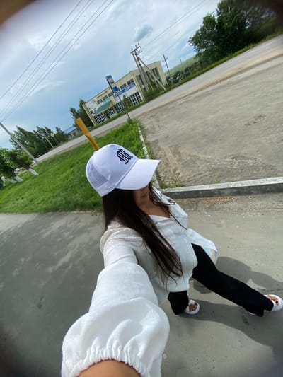 An outdoor, wide-angle, eye-level selfie is taken by a young woman wearing a white baseball cap with a black emblem, a white long-sleeved shirt, and black pants. She is standing on a paved area, possibly a sidewalk or road, with a curb to her right. In the background, a two-story building with a sign that reads "ТОРГОВЫЙ ДОМ" (Trading House) is visible, along with utility poles and trees. Beyond the building, a green, hilly landscape stretches under a cloudy sky. The scene appears to be in the city of Akyar, Russia, given the signage. The woman is holding the camera with her right hand, her arm extended.
