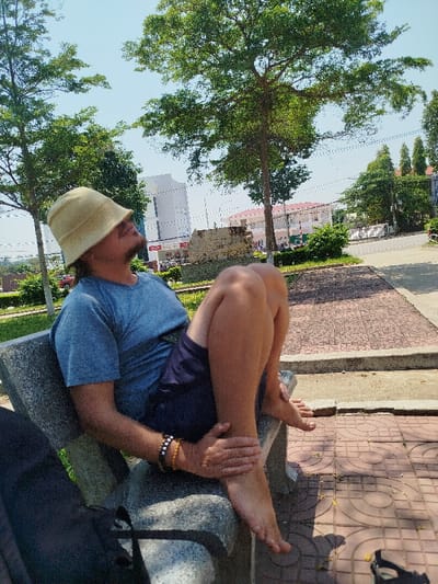 An adult male is seated on a light grey concrete bench in the city of Đăk Xú, Vietnam. He wears a light beige bucket hat, a blue t-shirt, and dark blue shorts. He has a prominent dark mustache and dark hair visible under his hat. His bare legs are bent, with his left hand, adorned with two beaded bracelets, resting on his right ankle. A black bag is partially visible on the bench to his left. The scene is outdoors under a bright, clear sky, indicated by strong shadows cast on the reddish-brown interlocking tiled pavement. Green trees frame the background, which includes a paved road, a multi-story white building, and a building with a red-tiled roof. A single black sandal rests on the tiled ground to the lower right.