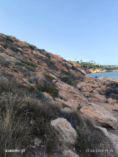 This media file depicts a scenic coastal landscape under a clear, bright blue sky, indicative of pleasant weather. The scene features a rocky, vegetated hillside sloping down towards the sea. The hillside is characterized by reddish-brown rocks, sparse dry grass, and some green shrubs. In the distance, along the coastline, there are buildings and palm trees, suggesting a developed coastal area.
The sea is calm and blue, meeting the rocky shore. On the shore, there are a few individuals visible. One person appears to be sitting on the rocks, while another is standing. The time of day is indicated by the timestamp to be late afternoon or early evening, specifically 19:15, and the lighting supports this, with a gentle, warm light illuminating the scene. The location is hinted at as Orihuela, Spain, which is known for its coastal areas and Mediterranean climate.
There is visible text in the lower portion of the image. In the bottom left corner, it reads "xiaomi 13T", which is likely a device identifier. In the bottom right corner, a date and time stamp reads "22.06.2026 19:15". The overall mood of the image is peaceful and serene, capturing a moment of quiet enjoyment of the coastal environment.