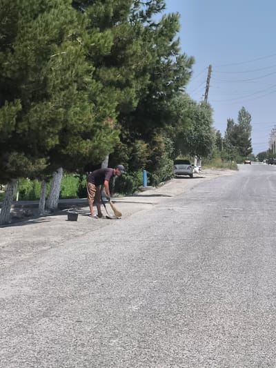 In Раджабгарди, Uzbekistan, a man wearing a dark t-shirt, brown shorts, a dark cap, and gloves is sweeping a dusty, paved road with a broom. A black bucket is placed on the ground next to him. The scene is outdoors with tall green trees lining the left side of the road. In the background, several cars are visible, including a white sedan parked on the side of the road and other vehicles further down the street. Power lines and a utility pole are also present in the background. The sky is clear and blue.