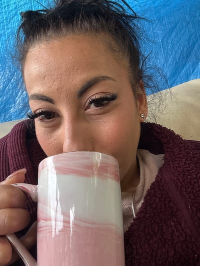 A woman lies on a light-colored surface covered with a blue sheet or cloth. She is wearing a burgundy-colored, textured, hooded garment over a light-colored top. She has dark hair and noticeable eyelash extensions. She holds a large white mug with pink marble swirl designs in front of her face with her right hand, where the manicured fingers with nail extensions are visible. The photograph does not contain location information about Warrington, United Kingdom.