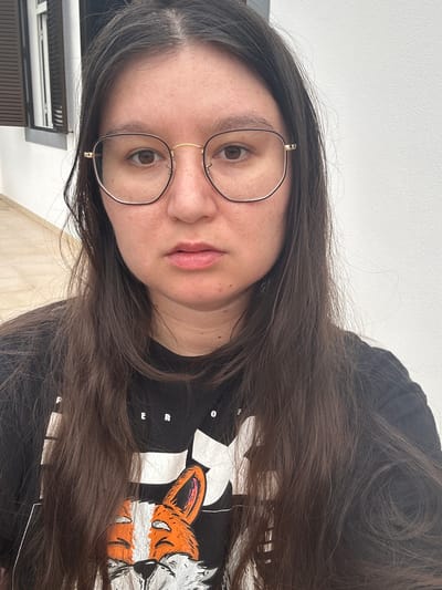 A woman with long brown hair and glasses is facing the camera. She is wearing a black T-shirt with a graphic design featuring a fox and lettering. The background includes a partially visible window with slats and a light-colored wall. The location appears to be indoors, possibly in Câmara de Lobos, Portugal.