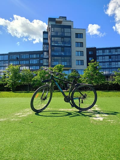 A black bicycle with turquoise accents is parked on a bright green artificial turf field. The bicycle has a front fender and a kickstand. In the background, a modern apartment building with many windows stands against a clear blue sky with scattered white clouds. Lush green trees and vegetation are visible between the field and the building. The scene appears to be outdoors on a sunny day, possibly in a residential area of Petrozavodsk, Russia. There are no people visible in the image.