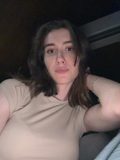 A medium shot shows a woman with shoulder-length brown hair and light skin from the chest up. She is looking at the camera with a neutral expression. Her eyes are brown, and she has a light makeup on her eyes and lips. She is wearing a light beige t-shirt and a silver bracelet on her right arm. Her left arm is bent upwards and resting on her head, and she is wearing a red string bracelet on her right wrist. The background is dark, with some lines visible at the top, suggesting an indoor setting with artificial lighting. The image was taken in Sharovechka, Ukraine.