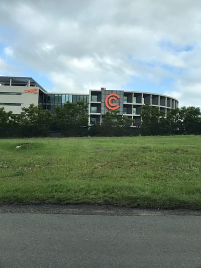 The image displays an exterior scene in Sandton, South Africa, featuring the Cell C head office building, a grassy field, and a section of asphalt road. The multi-story Cell C building dominates the background, characterized by light grey facades and extensive dark glass windows. An orange "cell C" logo is visible on the left, more angular section of the building, while a large, orange 'C' logo is prominently displayed on the curved right section, set against a dark grey panel with square apertures. Green trees partially obscure the building's lower levels. The midground consists of a broad, unmanicured green field, containing scattered small purple flowers and a white object, possibly debris, in the far-left area. The foreground shows a dark grey asphalt road, transitioning to a slightly elevated, darker strip of ground that borders the grassy field. The sky is overcast with grey and white clouds. No individuals, specific actions, or interactions are discernible.