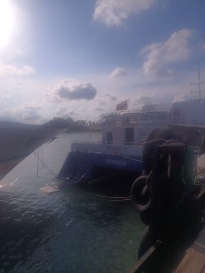 The image depicts a waterfront scene in Hat Sai Ri, Thailand, featuring a large ferry or boat docked alongside another vessel labeled "ROYAL 1 BANGKOK." The water is choppy. The sky is partly cloudy. A Thai flag is visible on the boat. There are trees in the background. The angle of the shot implies the photographer is on another vessel.