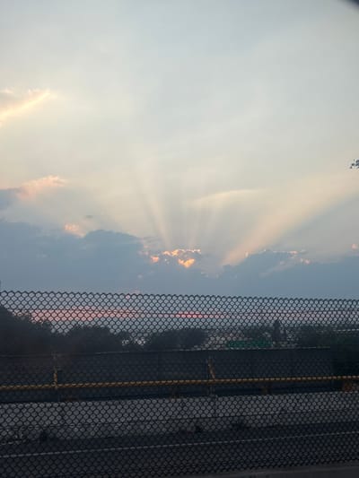 The image depicts an outdoor environment in Tultitlán, Mexico, observed during twilight hours, likely sunrise or sunset. The upper two-thirds of the frame feature a sky with a central cluster of dark and light clouds from which numerous bright, crepuscular light rays emanate and fan outward. The light source, partially obscured by these clouds, casts golden and white light, illuminating cloud edges with orange and yellow hues against a pale blue-grey upper sky. A dark, galvanized chain-link fence with diamond-shaped mesh dominates the lower third of the image, extending horizontally across the frame. Behind this fence, a dark, solid structure, likely a concrete barrier or wall, is visible. A single horizontal yellow pipe or rail is affixed to this dark structure, positioned approximately at the vertical center of the fence. Beyond these immediate structures, a diffuse landscape of dark tree canopies and faint, scattered urban lights is discernible towards the distant horizon. No individuals or specific actions are present within the scene.