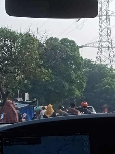 The image captures a street scene in Tangerang, Indonesia, viewed from inside a vehicle. Dominating the background is a large metal electricity pylon, characteristic of urban infrastructure. In the mid-ground, lush green trees line the street, partially obscuring a crowd of people. The people are diverse, with some wearing headscarves and others motorcycle helmets. One person is wearing a prominent orange helmet. Some individuals appear to be on motorcycles, suggesting traffic or a gathering. A blue tarp is visible, possibly covering goods or providing shelter. The foreground is filled by the dashboard of the vehicle, which includes a navigation screen displaying a map and indicating a distance of "3.6 km" to "Jl. Ciremai Raya," a street name common in the Tangerang region. The overall atmosphere is overcast, with a bright sky.