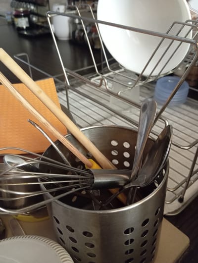 The image depicts a close-up view of kitchen items in a drying rack, likely in a domestic setting. The primary subjects are various kitchen utensils and containers. A white bowl is visible on the left, resting within the metal rack. To the right of the bowl, a blue plastic lid is partially seen. The central focus is a stainless steel utensil holder filled with various items, including a whisk, spoons, forks, and wooden chopsticks. An orange item, possibly a mat or a coaster, is placed beneath some of the utensils. The background shows the ribbed texture of a drying rack and a dark countertop. The lighting suggests an indoor environment, likely during the daytime, given the brightness. There are no people visible in the image, and no text is present. The overall scene conveys a sense of everyday domesticity, with items in the process of drying after being washed. The location context of Baden, Austria, suggests a European household setting.