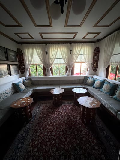 The image depicts the interior of a room, likely a living area, furnished in a traditional style. The room is dominated by a large, sectional sofa that wraps around the space, upholstered in a striped fabric with patterned cushions in shades of blue and cream. Several small, octagonal wooden tables with inlaid decorative patterns are placed in front of the sofa.
A patterned rug, primarily red with intricate white floral motifs, covers a significant portion of the floor. The room is illuminated by natural light streaming through large windows covered with sheer white curtains. The windows offer a view of lush green foliage, suggesting an outdoor garden or natural landscape.
The ceiling features a decorative wooden trim creating rectangular panels. On the walls, there are framed black and white photographs and ornate wall hangings. The overall ambiance is one of comfort and traditional elegance.
This scene is located in Eyüpsultan, Turkey, within a historical building that appears to be a traditional Ottoman-era residence or a meticulously preserved historical space. The architectural details, furniture style, and decorative elements are characteristic of the region's rich cultural heritage.