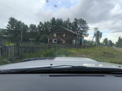 The image shows a view from inside a vehicle, looking out at a wooden house in the city of Gremiuchiy, Russia. The house is two stories tall with a dark wooden exterior and multiple windows. A wooden fence runs in front of the house, and there are trees and a dirt road in the background. The sky is cloudy.