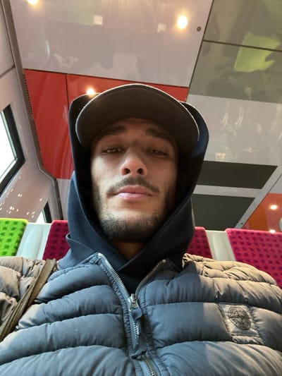 An image depicts a male individual taking a selfie inside a modern passenger train, likely in the city of Bois-Colombes, France. The subject, a man estimated to be between 20 and 35 years old with dark hair and a beard, looks directly into the camera with a neutral expression. He is wearing a dark baseball cap, a dark hooded sweatshirt with the hood pulled over the cap, and a charcoal grey puffer jacket. A circular patch logo, identified as the Stone Island compass, is visible on the upper left sleeve of his jacket. A dark strap from a bag is seen over his right shoulder. The train's interior features light-colored, possibly reflective, ceiling panels with embedded warm-toned circular light fixtures. Seating in the background exhibits vibrant patterned upholstery in alternating green and magenta/red squares. A window on the left side of the frame shows a blurred exterior, and reddish-orange wall paneling is visible on the right.