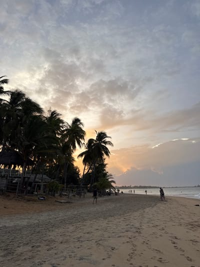The image depicts a wide sandy beach in Arugam Bay, Sri Lanka, during what appears to be sunset. The sky is filled with dramatic clouds, tinged with orange and yellow hues from the setting sun, which is partially obscured by clouds on the right side of the frame. Palm trees line the edge of the beach on the left, casting shadows and framing the scene. In the background, several thatched-roof structures and wooden walkways suggest a resort or beachside establishment.
Several people are visible on the beach. In the mid-ground, a few individuals are scattered along the shoreline, some walking, others standing. On the right side of the beach, a couple is embracing, facing the ocean. Further down the beach, more figures can be seen, some possibly swimming or wading in the water. The sand is marked with numerous footprints, indicating recent activity. The ocean waves are gentle, rolling towards the shore. The overall atmosphere is serene and tropical, characteristic of a beach destination at dusk.
