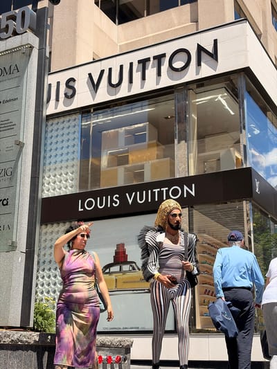 This media file is a vertically oriented, full-body photograph taken outdoors in what appears to be a city setting, likely Toronto, Canada, given the context provided. The primary subjects are two individuals standing in front of a Louis Vuitton store.
The person in the foreground on the right is wearing a tie-dye dress in shades of purple, pink, and green. They are wearing sunglasses and have their hand on their head, possibly posing or shielding their eyes from the sun. Their expression is not clearly discernible.
The second person, positioned to the left and slightly behind, is dressed in a black and white striped full-body outfit, including a dramatic ruffled collar. They are also wearing a golden headpiece and sunglasses, and are holding a smartphone in their right hand. Their pose suggests they might be looking at their phone or interacting with it. A man in a light blue shirt and dark pants with a blue baseball cap is visible in the background, to the right of the second individual.
The background prominently features the facade of a Louis Vuitton store, with its distinctive "LOUIS VUITTON" signage visible. The store appears to have a large display window. Also visible are some lower-level signs with text, including "OMA," "Edelman," and "ZIG INSTITUTE." The overall lighting suggests it is daytime, likely sunny, casting shadows on the ground and the building. The scene appears to be a street view in front of a high-end retail establishment.