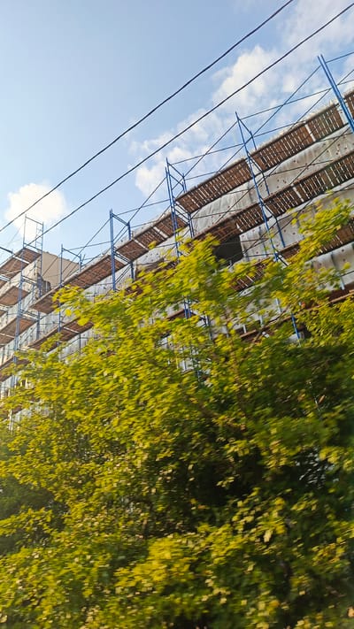 The image captures a multi-story building undergoing renovation, shrouded in scaffolding. The scaffolding consists of a blue metal frame with wooden planks serving as platforms. The building itself appears to be covered in a white insulating material, with visible wooden beams or supports. In the foreground, lush green foliage from trees partially obscures the lower sections of the building and scaffolding. The trees are blurred, suggesting movement, possibly from a passing vehicle.
The sky above is a clear blue with scattered white clouds. Two dark electrical wires stretch diagonally across the upper portion of the frame. The lighting indicates it is daytime, likely late afternoon or early morning, given the angle of the sun and the shadows cast. There are no people visible in the image. The scene suggests an ongoing construction project in an urban or semi-urban setting, with the presence of mature trees indicating a developed area. The overall impression is one of progress and development.