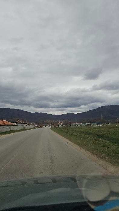 The image displays a scene in Kliment, Bulgaria, captured from within a vehicle, indicated by a reflection on the dashboard at the bottom. A grey asphalt road occupies the foreground and midground, extending towards a distant settlement. To the immediate left of the road, a low concrete wall precedes several residential-style buildings with light-colored facades and red-tiled roofs, some appearing unfinished. To the right, an expanse of green grass and dirt constitutes undeveloped land. Further along the road, a cluster of more distant buildings is visible, one of which prominently features a slender minaret with a conical cap. A large, dark-colored truck is observed moving away from the viewer on the road in the midground. The background consists of dark, undulating mountains or hills, covered with sparse vegetation. The sky is uniformly overcast with heavy, grey clouds, indicating dull or impending weather. No individuals are discernible.