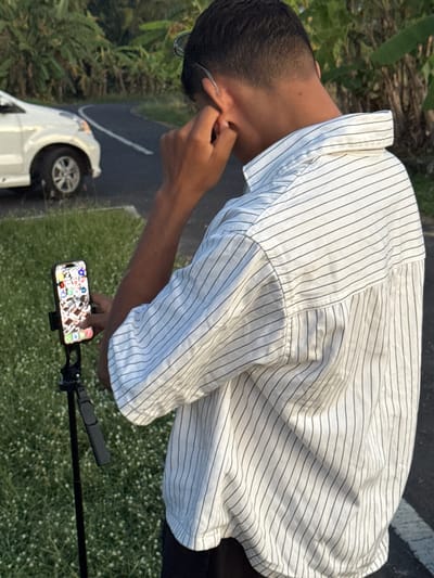 A young man with short dark hair is adjusting his eyeglasses, dressed in a vertically striped white shirt, standing outdoors in Selemadeg Timur, Indonesia. A white car is parked on the asphalt nearby, and a smartphone is mounted on a stand in front of him, on a patch of grassy land.
