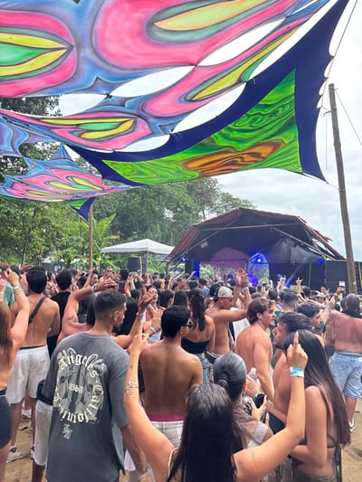This image depicts an outdoor festival or event in Cairu, Brazil, under an overcast sky. Overhead, multiple large, abstractly patterned fabric canopies in vibrant shades of pink, blue, green, yellow, and orange are suspended, with some sections featuring solid dark blue fabric.

In the mid-ground, a makeshift stage with a dark, gabled roof is prominent, displaying blue artificial lighting and speakers on its sides. To the left of the stage, a smaller white square canopy tent is visible. The background consists of dense green tropical foliage.

The foreground and mid-ground are densely populated by a crowd of approximately 50-100 individuals, predominantly young adults. Most attendees are facing the stage, with many raising their hands, indicative of dancing or cheering. Attire is casual, including shirtless men, t-shirts, tank tops, and shorts. Several individuals wear event wristbands. A person in the central foreground is seen from behind wearing a dark grey t-shirt with a white "Angeles USA" graphic. Another individual in the right foreground has a spiral-design gold-toned armlet. The ground beneath the crowd is unpaved dirt.