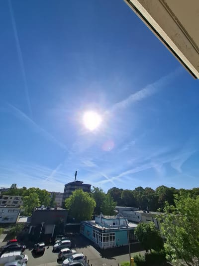 The image captures an outdoor scene looking upwards towards a bright sun in a clear blue sky. Multiple contrails from aircraft are visible, crisscrossing the sky. Below the sky, an urban environment is depicted, featuring buildings of various heights and architectural styles, including apartment buildings, a taller commercial building with an antenna on top, and a smaller building with a sign that appears to read "King's Diner" and lists food items like "Doner Pizza Burger." Several cars are parked in a lot in front of these buildings. Lush green trees are interspersed throughout the urban landscape. The presence of the "King's Diner" and the general urban architecture suggests the location is likely in a European city, potentially Trier, Germany, based on the context provided. The time appears to be daytime, given the strong sunlight and clear sky. No people are distinctly visible or engaged in specific actions, though a small figure might be present near the diner. The overall impression is a sunny day in a city.