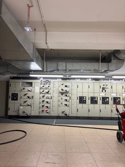 This image depicts an interior view of an electrical control room, likely located in a commercial or industrial facility within the city of Mannheim, Germany. Dominating the scene is a large, modular electrical panel system extending horizontally across the frame. The panels are light gray with numerous compartments, each featuring black levers, switches, and indicator lights, suggesting active electrical equipment. Some compartments have labels, though they are largely illegible from this distance. Above the panels, industrial ventilation ducts made of galvanized metal are prominently installed on the ceiling, along with fluorescent lighting fixtures. In the foreground, a black electrical cable is coiled on the tiled floor. To the far right, a red fire extinguisher on wheels is partially visible. The overall environment is utilitarian and functional, characteristic of a power distribution or control center.