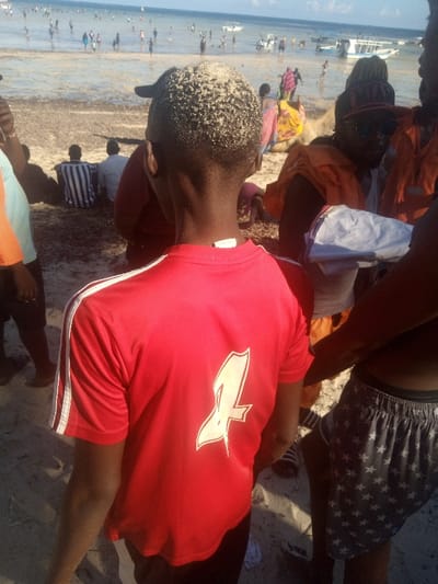 The image displays a densely populated beach scene in Shanzu ward, Kenya, under a clear daylight sky.
In the immediate foreground, the back of a young individual is visible, characterized by short, dark hair partially coated with sand. They are wearing a red short-sleeved t-shirt featuring two white stripes on each shoulder/sleeve and a large white "4" emblem on the back. To the right, the lower body of another individual is partially visible, wearing dark shorts patterned with white stars.
The midground features several people and objects. A man with a beard, sunglasses, and a backward baseball cap is visible, wearing an orange garment draped over a white tank top, and holding a folded light-colored cloth. Adjacent to him, a camel stands, draped with a multi-colored blanket in yellow, pink, and red hues. Further back on the sand, two individuals are seated, one in a black and white striped shirt, the other in a light-colored shirt. Patches of dark seaweed are scattered across the sandy shore.
The background depicts the ocean, populated by numerous individuals wading, standing, and swimming in the shallow water. Several small boats are visible further offshore against a bright blue sky with minimal cloud cover.