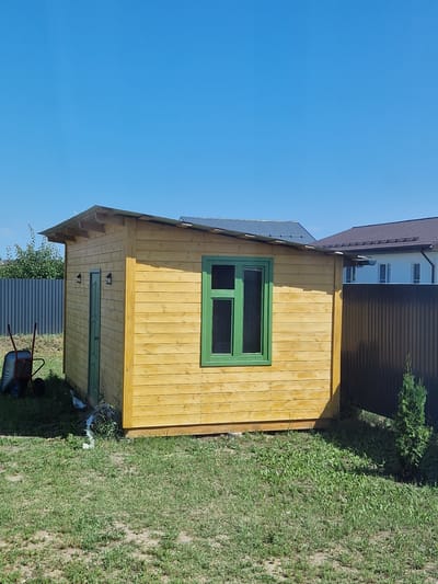 A small wooden shed with a green window and door is situated in the foreground, bathed in sunlight under a clear blue sky. The shed's exterior is constructed from horizontal wooden planks with a light yellow-brown finish. A metal roof with a visible slope extends outwards. To the left of the shed, a wheelbarrow rests on the grass, and a gray corrugated metal fence runs along the property line. In the background, neighboring houses with tiled roofs are visible, along with a dark brown corrugated metal fence. The ground is covered with green grass, some of which appears dry and patchy. The scene is likely in a residential area on a sunny day.