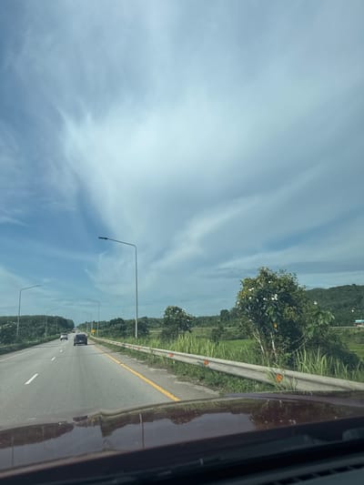 The image is a photograph taken from the perspective of someone driving on a highway in Tambon Khao Tok, Thailand. The highway has multiple lanes and is bordered by a guardrail and lush green vegetation, including trees and tall grass. In the distance, rolling hills covered in trees are visible. The sky is a prominent feature, with dramatic, wispy white clouds against a blue backdrop. Several streetlights line the side of the road. Cars are visible on the highway, indicating it is in use. The foreground shows the hood of the vehicle from which the photo is taken, reflecting the sky and surrounding landscape. The overall impression is that of a scenic drive through a rural or semi-rural area.
