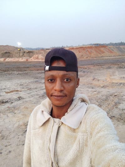 A man in a dark baseball cap and light-colored, textured jacket stands in a dry, dusty landscape. Behind him, a series of mounds and a structure, possibly a building, are visible. In the distance, a bright light source illuminates the scene. The man is facing the camera with a neutral expression. The setting appears to be an industrial or construction site.