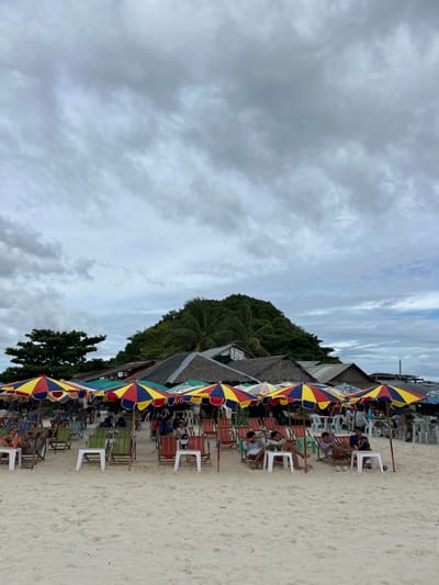 This image depicts a beach scene in Phru Nai, Thailand, on a cloudy day, likely in the late afternoon. The foreground is dominated by a sandy beach with numerous rows of beach chairs and colorful umbrellas, predominantly red, yellow, and blue. Several people are seated in the chairs, some appearing relaxed and others engaged in conversation. The umbrellas provide shade, suggesting a break from intense sun, which aligns with the cloudy weather.
Behind the chairs, there are small thatched-roof structures that appear to be beachside vendors or food stalls. Further back, a lush, green hillside covered in dense trees, including palm trees, rises steeply. The sky is filled with dramatic, layered clouds, some dark and heavy, indicating potential for rain, while others are lighter, allowing diffused sunlight to filter through. The overall mood is one of leisure and relaxation, typical of a beach resort setting. There is no visible text in the image.