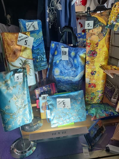 The image displays a retail setting, likely a shop in Thessaloniki, Greece, showcasing a variety of artistic merchandise. The items are primarily bags and wallets featuring famous artworks. Dominating the display are bags and accessories adorned with Vincent van Gogh's "Starry Night" and "Almond Blossoms," as well as Gustav Klimt's "The Kiss."

Several items are tagged with prices in Euros, indicated by a "€" symbol and accompanying numbers. For instance, a bag with a Van Gogh print has a "5" followed by a comma and ".€", suggesting a price of 5 Euros. Another "Starry Night" item is marked with "10.€", indicating a price of 10 Euros. A clutch featuring Klimt's "The Kiss" also displays a "5.€" tag. Another item, a wallet, shows "8.€" implying a price of 8 Euros.

The merchandise is arranged on shelves and stands, with some items hanging from a rack. The background is dimly lit and suggests a store interior, with racks of clothing and other retail fixtures visible. The overall impression is a display of art-themed consumer goods.
