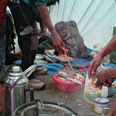 This image captures an active outdoor food preparation scene at Gilman's Point, Tanzania. The environment is a makeshift camp kitchen, indicated by a white tarp serving as a backdrop and a blue tarp spread on the muddy ground. Several individuals are present, engaged in meal preparation. A person wearing black and grey athletic pants and brown hiking shoes is partially visible on the left. In the center, a person's hands are seen holding an orange vegetable peeler over a piece of produce, possibly a potato, with steam or vapor rising from the area. A red plastic bowl on the blue tarp contains numerous vegetable peels and a large kitchen knife. Fresh cucumbers and red tomatoes are also visible on the blue tarp near the bowl. To the right, another person's hands are holding a yellow bottle of "Sunola Sunflower Oil" and a blue tub of "Blue Band" margarine. A metal gas burner ring is visible in the foreground, and a large silver thermos or kettle with a white lid and a pair of metal tongs is positioned beside it. Stacked metal plates or pot lids are on the blue tarp to the left of the red bowl. A dark, large backpack is leaning against the white tarp in the background, near a green bag.