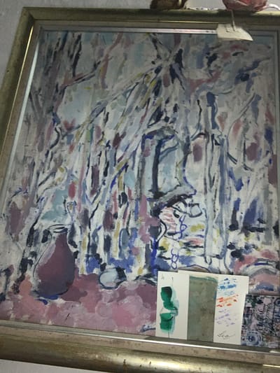 A framed abstract painting is displayed. The painting features broad brushstrokes in shades of white, blue, pink, and purple, creating a somewhat chaotic yet harmonious composition. In the foreground, at the bottom right, a small collection of cards or postcards are placed. One card has three distinct sections: a green abstract figure on the left, a textured gray rectangle in the middle, and a section with blue and orange abstract marks above purple smudges on the right, with a handwritten signature "Leo" below it. To the right of this, another small card with a complex pattern is visible. The painting is housed in a metallic gold-colored frame. The backdrop appears to be a wall. The scene is indoors. No specific location in Tacarigua, Venezuela, can be determined from the image.