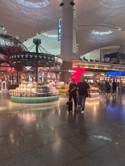 This is a full shot of the interior of an airport terminal in Arnavutköy, Turkey. The scene is dominated by a modern, high-ceilinged space with a slatted white ceiling structure. In the foreground, a couple walks away from the camera towards the left. The man is in the center, wearing a dark t-shirt and blue jeans, and is pulling a black suitcase. To his left, a woman in a dark t-shirt and pants walks beside him, also pulling a suitcase.
In the mid-ground, a circular display stand is filled with boxes of Turkish delight and baklava. Behind this, a neon sign reads "TURKISH" and "BAKLAVA". Further back, there are various shops and seating areas visible. People can be seen walking and sitting in these areas. Blue directional signs indicate "Kapı / Gate". The floor is polished dark stone, reflecting the overhead lights and the displays. The overall atmosphere is busy and commercial, typical of a large international airport.